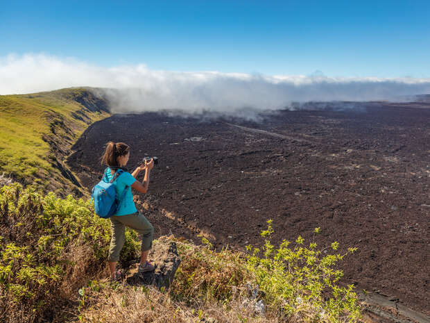 A person stands on a grassy cliff, photographing a vast, dark volcanic landscape stretching beneath a clear blue sky with scattered clouds.