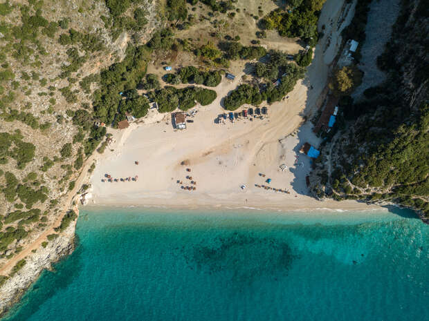 A sandy beach with scattered umbrellas and chairs rests between turquoise water and rocky cliffs with green foliage. Small structures and trees are situated behind the beach, creating a tranquil atmosphere.