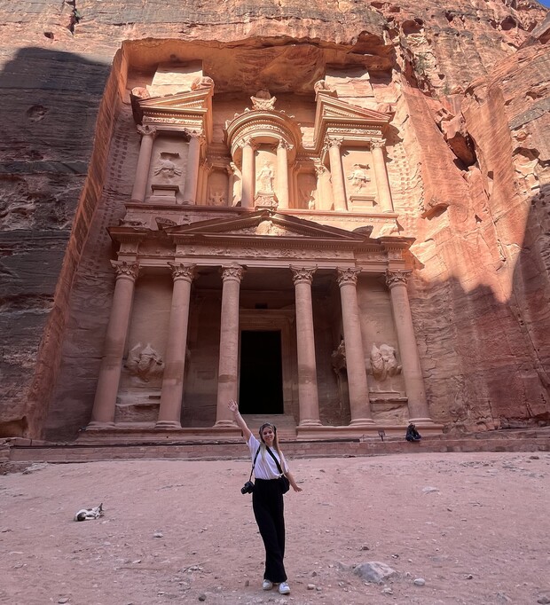 A woman stands in front of Petra's Treasury, a monumental rock-cut facade with columns and intricate carvings, surrounded by towering reddish cliffs in the ancient city.