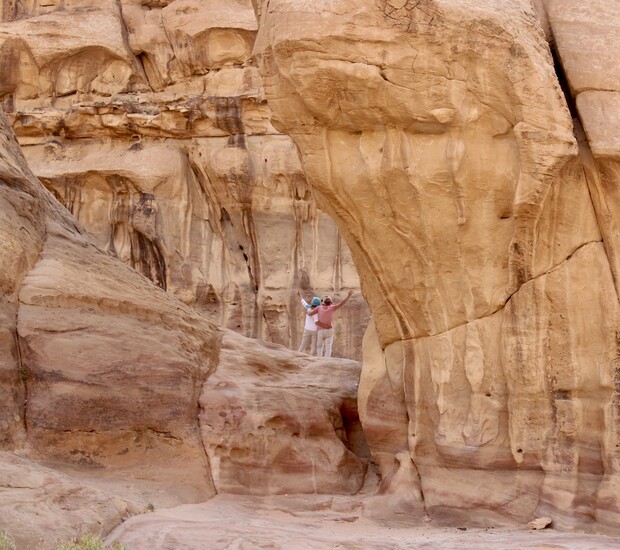 Two people stand amidst towering sandstone formations, arms outstretched, in a rugged, desert canyon setting with layered rock textures and soft, warm colors.