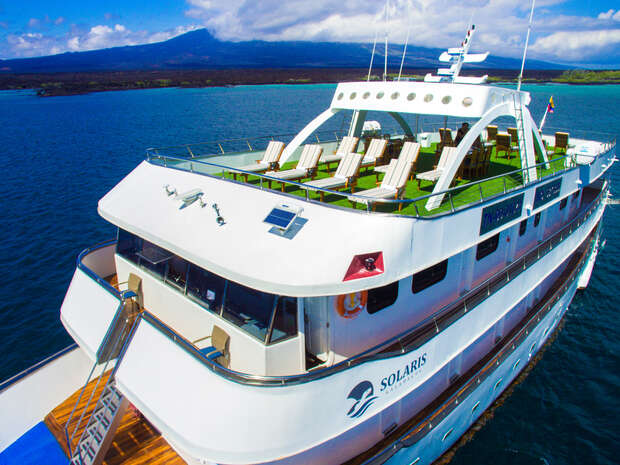 A large white yacht with "SOLARIS" written on the side features a deck with lounge chairs. It is docked on calm blue water, with distant mountains and a partly cloudy sky.