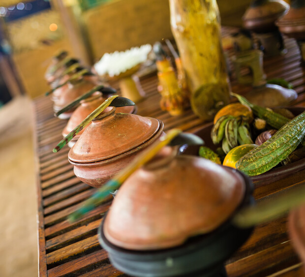 Clay pots with ladles rest on a wooden table, surrounded by various vegetables and bottles, in a rustic kitchen setting.