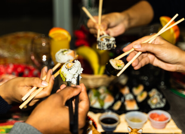 Hands holding chopsticks lift sushi pieces over a table filled with assorted sushi, sauces, and fruit. Glasses with orange slices and a basket of bananas are visible in the background.