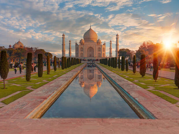 Taj Mahal stands reflected in a long rectangular pool, surrounded by lush gardens and tall slender trees under a vibrant sky during sunset. Visitors walk along the pathways.