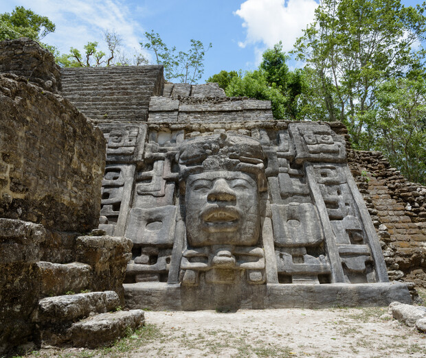 Stone mask adorns ancient Mayan temple, surrounded by stair-like ruins, set in lush, wooded jungle. Bright sky above enhances the contrast between nature and man-made structure.