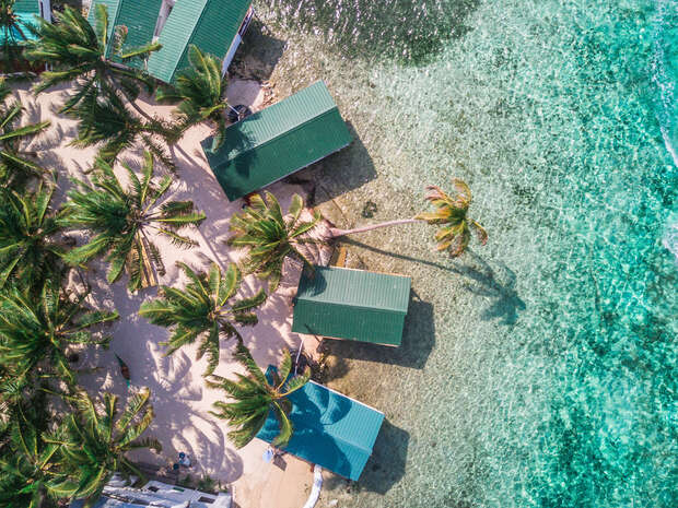 Green-roofed huts stand amid palm trees on a sandy beach, bordering clear, shallow waters. The scene is viewed from above, showing the lush, tropical environment and the vibrant coastline.