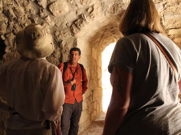 A person in an orange shirt speaks to two people in a stone archway, suggesting a historical or archaeological context. Light enters from a doorway behind them.
