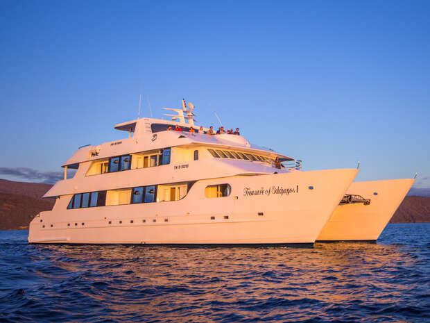 A large white yacht labeled "Treasure of Galapagos" floats on calm water during sunset. People are gathered on the upper deck, surrounded by distant mountains and a clear sky.