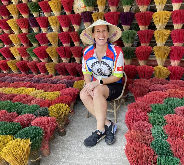 A person wearing a conical hat sits smiling on a wooden chair, surrounded by colorful bundles of incense sticks arranged in neat patterns against a wall.