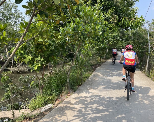 Cyclists ride along a narrow, shaded concrete path surrounded by lush greenery and a small waterway, under a clear blue sky.