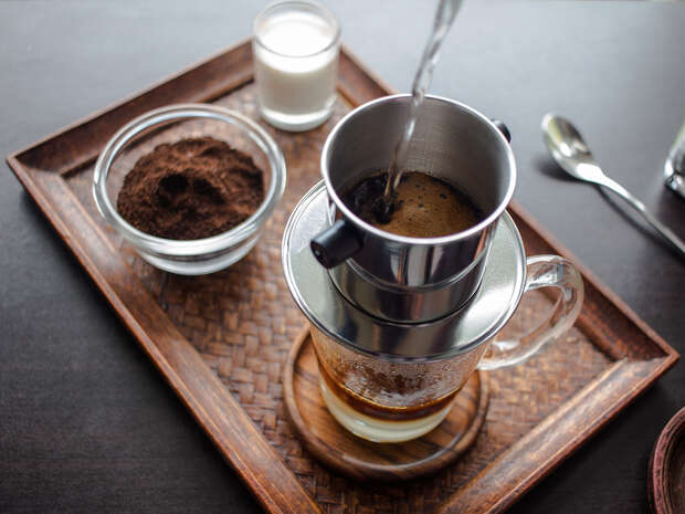 A stainless steel coffee filter drips coffee into a glass mug on a wicker tray, surrounded by a bowl of ground coffee, a small glass of milk, and a spoon.