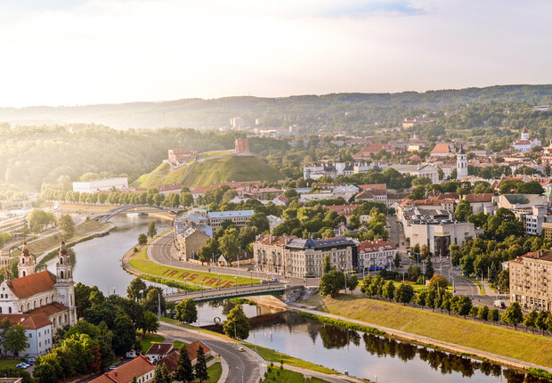 Arial view of a city with a river curving through, historic buildings, and green hills. A castle is visible atop a hill. Text on riverbank reads "IR AS TU!"