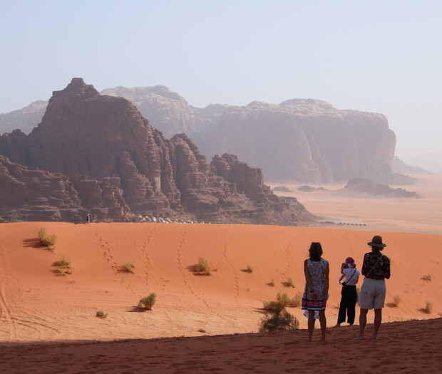 Three people stand on orange desert sand, observing the large sandstone rock formations in the distance. Sparse bushes dot the landscape under a hazy sky.