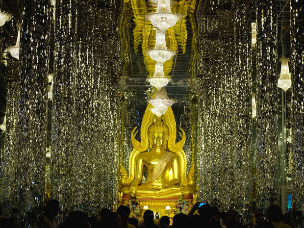 A large golden Buddha statue sits serenely amidst hanging crystal strands, reflecting light. Overhead, chandeliers illuminate the space. Dark silhouettes of people are visible in the foreground.
