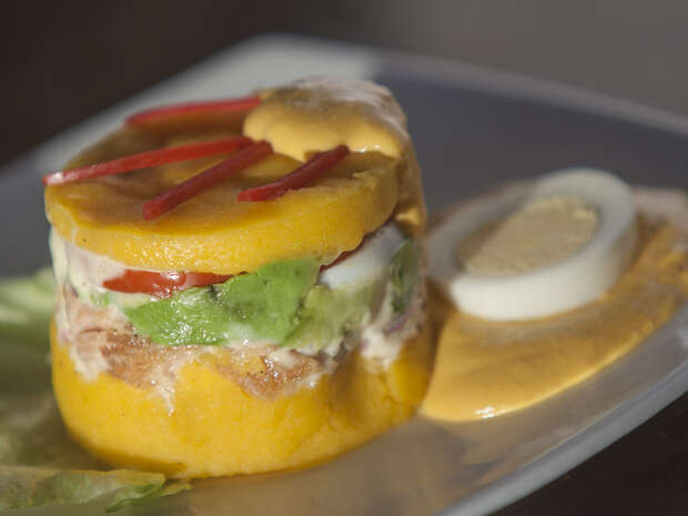 A layered Peruvian causa showcases mashed yellow potatoes, avocado, and sliced vegetables, served with creamy sauce on a white plate alongside sliced boiled egg and lettuce.