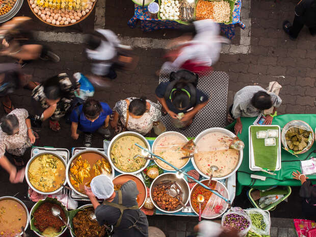 Crowded street market scene with food vendor serving colorful dishes from large pots. People move around, some standing in line, surrounded by vibrant produce and a bustling atmosphere.
