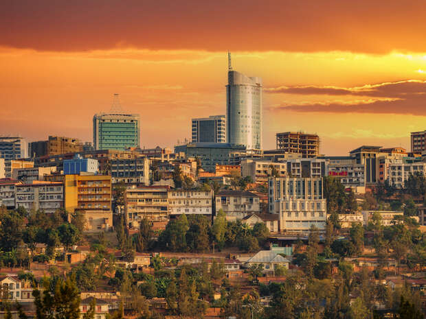 A skyline featuring tall buildings rises against a vibrant orange sunset, with scattered trees and lower structures in the foreground, creating an urban landscape at dusk.