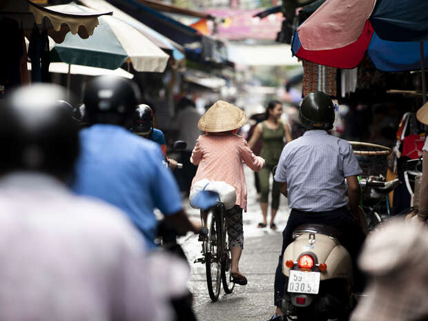 A cyclist wearing a conical hat rides through a bustling, narrow market street, surrounded by people on motorcycles and pedestrians under colorful awnings.