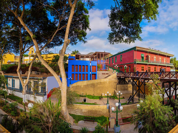Colorful buildings stand amidst lush trees, framing a wooden bridge. The scene captures a vibrant neighborhood, offering a mix of nature and architecture under a bright, blue sky.