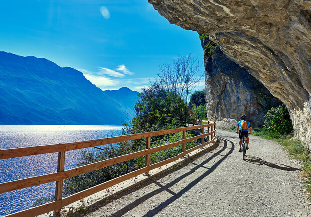 Cyclist rides along a gravel path bordered by a wooden fence, passing through rocky cliffs. The path overlooks a bright blue lake with mountainous scenery under a clear sky.