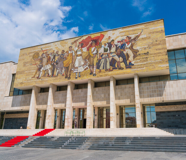 Mosaic mural depicting historical figures in action, marching forward with determined expressions. Positioned above the entrance of a large stone building featuring columns and a red carpeted staircase.