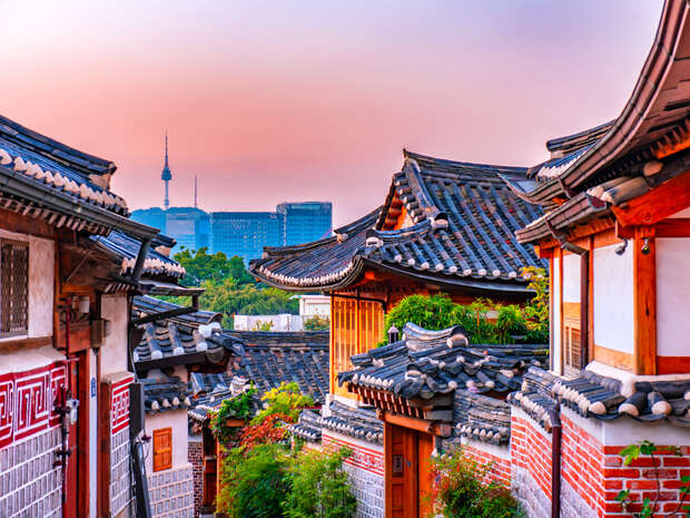Traditional Korean village rooftops rest under a vibrant sunset sky. In the background, a modern cityscape with tall buildings and a tower contrasts with the historical architecture.