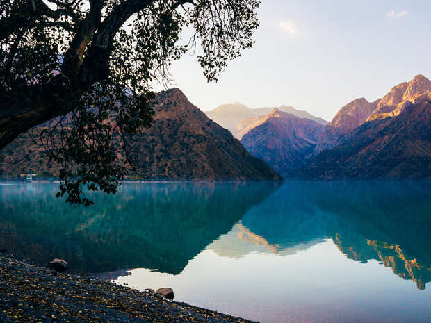 A tree extends over a calm lake reflecting surrounding mountains, under a clear sky. Sunlight softly illuminates the rugged peaks, enhancing the tranquil, scenic landscape.