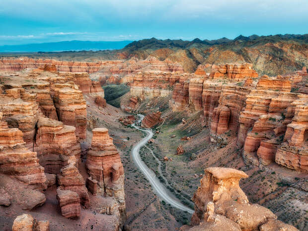 Canyon with towering reddish-brown rock formations lines a narrow, winding dirt road cutting through the valley. In the distance, mountains rise under a vast, partly cloudy sky.