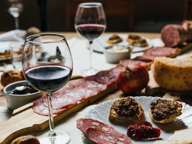 Wine glasses are filled with red wine, surrounded by a charcuterie spread on a wooden board. Various meats, cheese, and small bowls of condiments are arranged on a dining table.