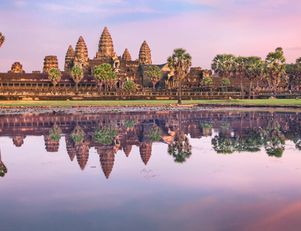 Ancient stone temple towers stand majestically under a pink-purple sky, reflected in a tranquil lake surrounded by palm trees and greenery, depicting serene, timeless beauty.