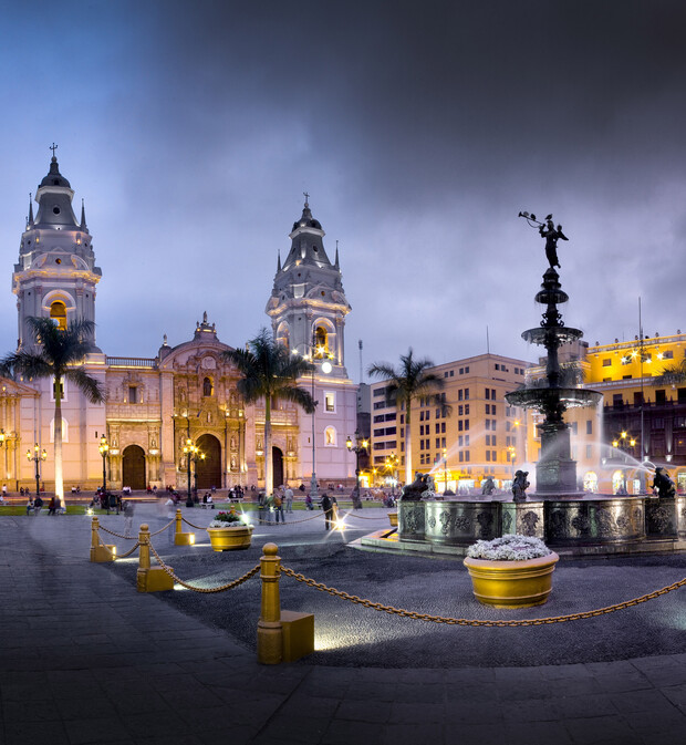 A fountain sprays water under cloudy evening skies in a brightly lit plaza, surrounded by yellow planters, palm trees, and an ornate cathedral in the background, with city buildings nearby.