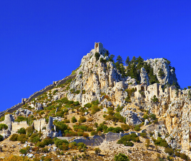 A rocky hilltop features ancient fortress ruins amidst sparse vegetation under a deep blue sky. Towering stone structures and walls dominate the landscape, surrounded by scattered trees.