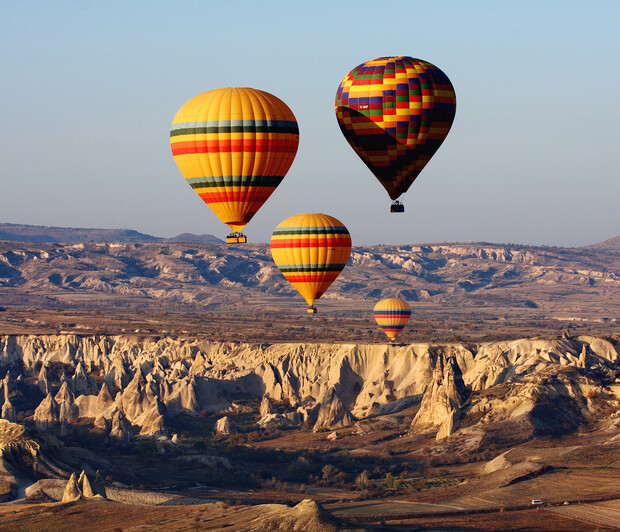 Colorful hot air balloons ascend over a rugged, rocky landscape with steep cliffs, under a clear blue sky, suggesting a scenic aerial journey.