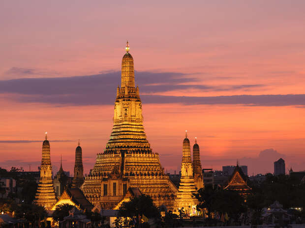 A majestic, illuminated temple with a tall spire stands at sunset, surrounded by smaller structures, silhouetted against a vibrant orange and pink sky, creating a serene and picturesque scene.