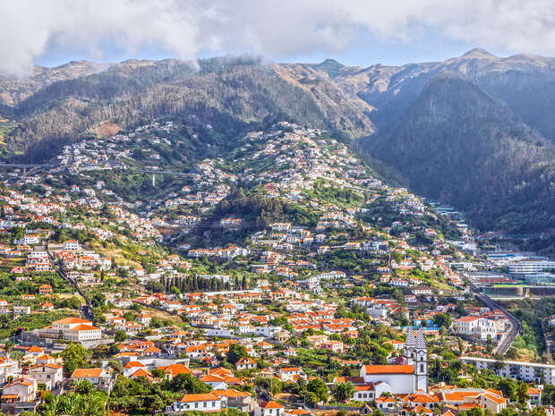 A sprawling town with red-roofed buildings is nestled on a lush, green mountainside. Roads wind through the settlement, surrounded by verdant hills under a partly cloudy blue sky.