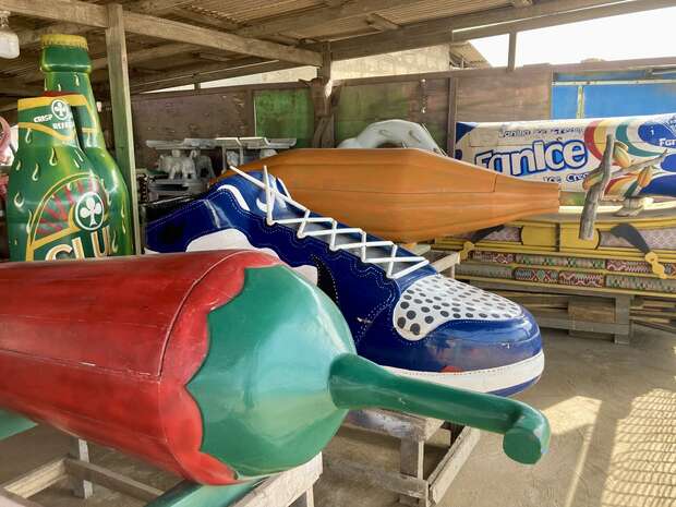 Colorful, oversized objects resembling a sneaker, bottle, chili pepper, and ice cream are displayed in a workshop-like setting. Wooden beams and various tools can be seen in the background.
