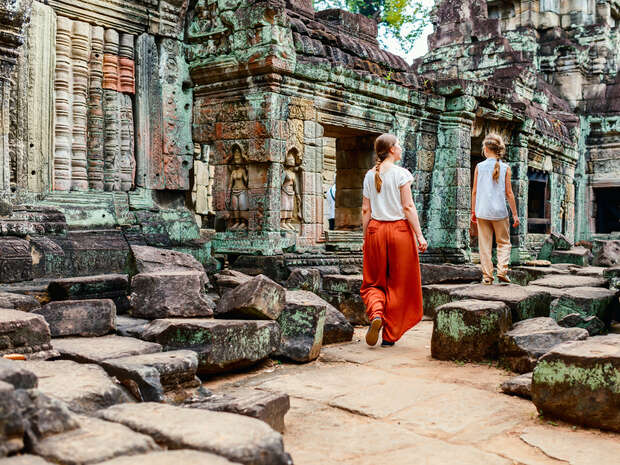 Two people explore ancient stone ruins with intricate carvings, surrounded by moss-covered structures and scattered stone blocks, under a tree-dappled sky.