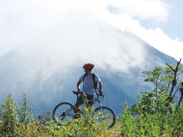 A cyclist stands with a bike on a grassy hill, overlooking a mist-shrouded mountain in the background, under a partly cloudy sky.
