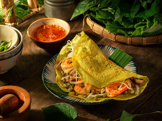 A Vietnamese pancake, filled with shrimp, pork, and bean sprouts, rests on a plate surrounded by leafy greens, a bowl of spicy dipping sauce, and traditional wooden utensils.