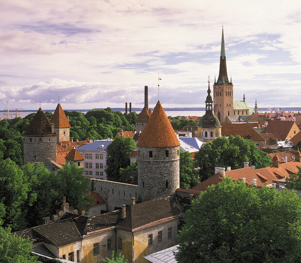 Stone towers with red roofs stand prominently amid a mix of historic and modern buildings, surrounded by lush greenery and under a partly cloudy sky, in a scenic urban landscape.