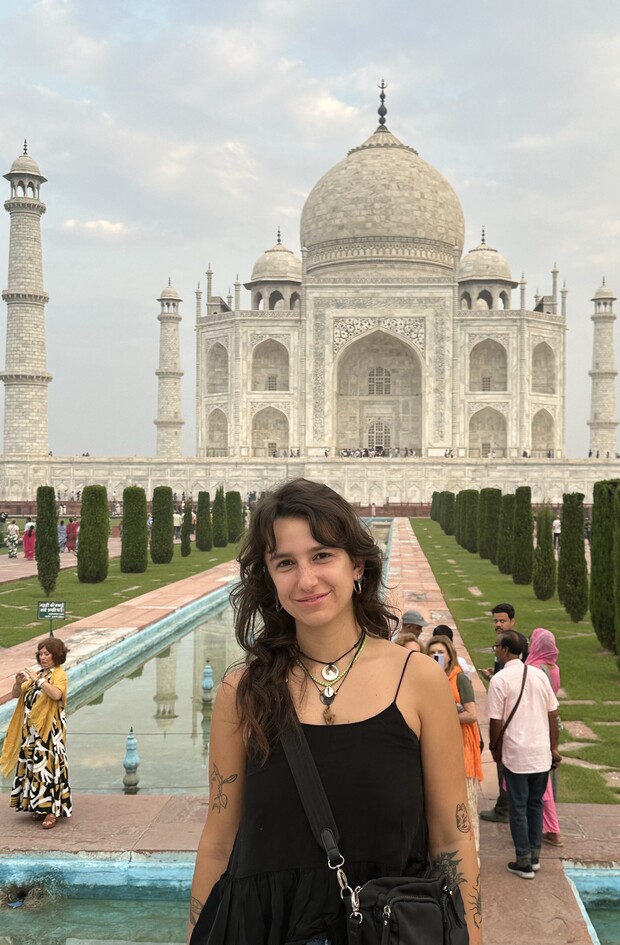 A person stands smiling in front of the Taj Mahal, surrounded by tourists and lush gardens with a reflecting pool leading towards the iconic white marble mausoleum under a partly cloudy sky.