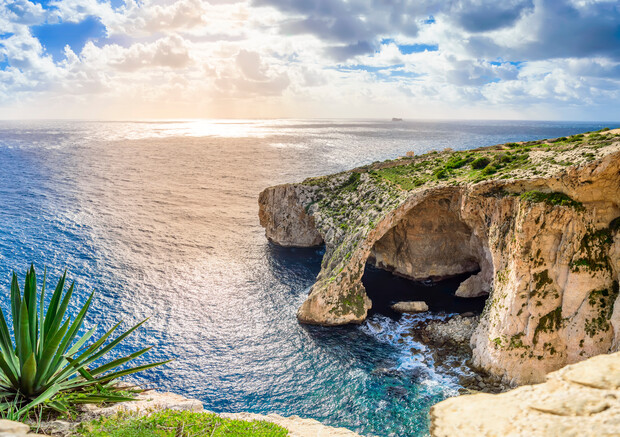 A coastal rock arch rises dramatically from a cliff, with waves crashing below in clear blue sea; lush greenery clings to the cliff, while the horizon glows under a cloudy sky.