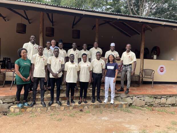 A group of fifteen people, wearing casual uniforms, pose together in front of a patio with wooden posts. They stand on a stone-paved area under a roof, surrounded by a natural outdoor setting.