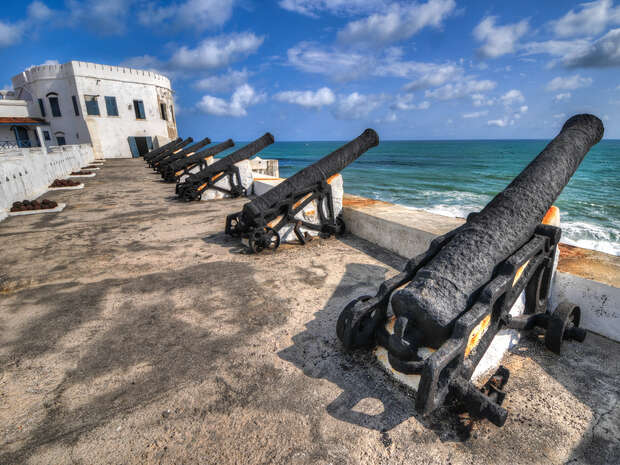 Cannons, aligned on a stone fortification, point towards the ocean under a partly cloudy sky. A white building with a weathered exterior stands nearby, enhancing the historical ambiance.