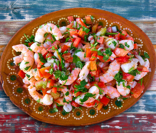 Shrimp is mixed with diced tomatoes, onions, and cilantro, garnished with herbs on a decorative, oval plate. The background consists of rustic, multicolored wooden planks.