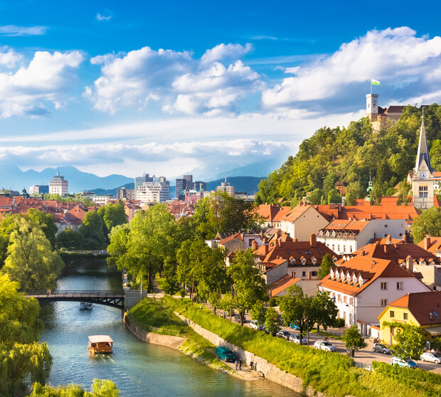 A river flows through a city with a boat traveling under a bridge. Surrounded by trees and red-roofed buildings, a hill with a castle overlooks the urban landscape.