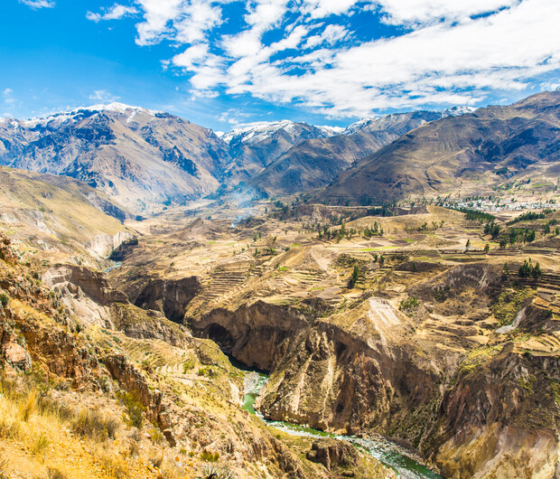 A deep canyon with terraced fields descends towards a flowing river, surrounded by rugged, snow-capped mountains under a partly cloudy sky.