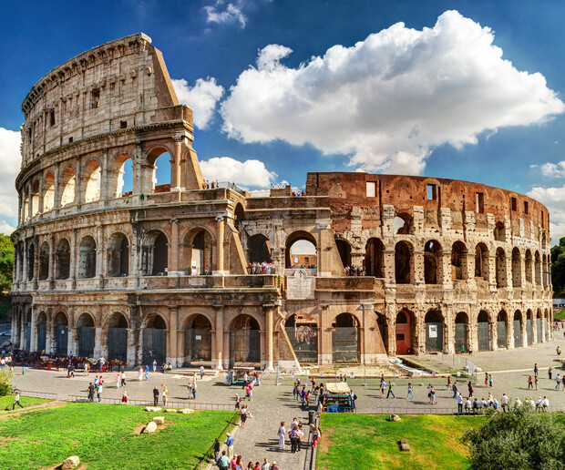 The Colosseum stands partially ruined under a bright blue sky with fluffy clouds, surrounded by tourists walking on a paved area, with patches of green grass nearby.