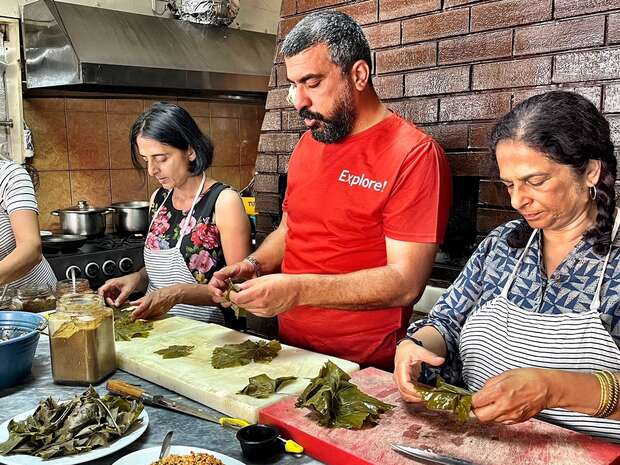 Three people prepare food by wrapping ingredients in leaves on a counter in a kitchen setting. Pots are visible on the stove in the background. One person wears a red shirt reading "Explore!".