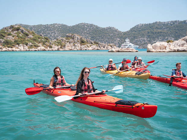 Kayakers paddle in vibrant red and yellow kayaks on clear turquoise waters. They are wearing life vests amidst a backdrop of rocky hills and a distant yacht.